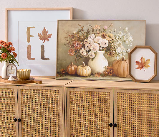 A wooden hallway table is decorated with affordable fall decor from pOpshelf including a small vase with orange fall flowers and a seasonal candle on a leaf serving tray and fall wall art with leaves, pumpkins, and autumn florals rested against the wall.