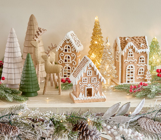 Ceramic gingerbread house decor in three styles on a table with wooden deer and ceramic trees surrounded by Christmas floral, berries & greenery.