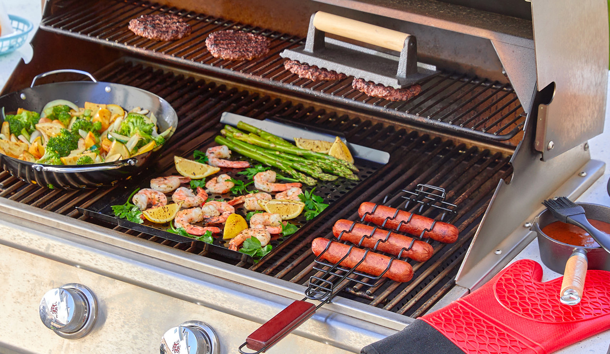 Hot dog griller, hamburger press, and flat and round grill pans on a grill surrounded by other grill tools and accessories.