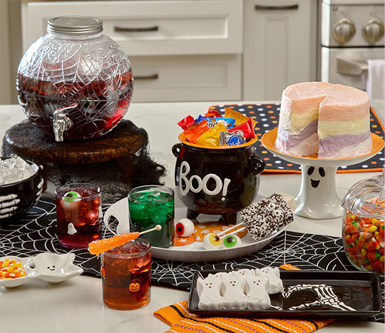 A kitchen island counter is decorated with classic Halloween-themed decor including a Boo! caldron, ghost cake display, spider web table runner, skeleton hand plates and bowls, a spider web Halloween drink dispenser, and Halloween icon glassware.