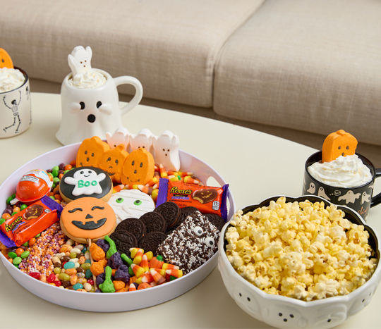Halloween candy & cookie charcuterie on a white round tray with Halloween mugs, popcorn in a ghost bowl, Halloween Peeps, and cocoa bombs.