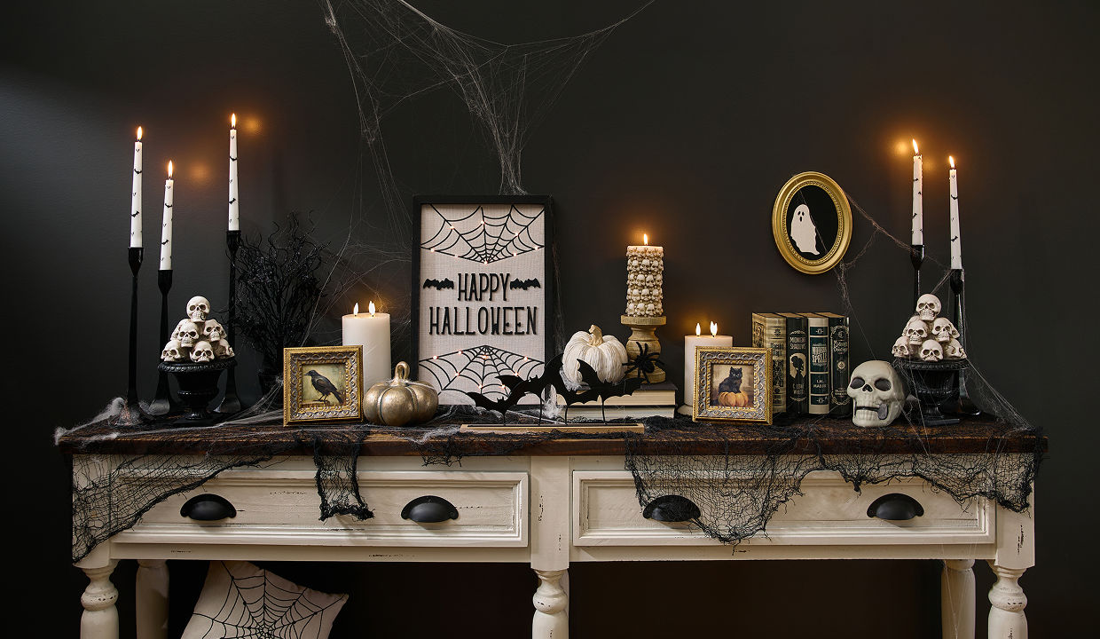 A spooky Halloween-themed foyer table is decorated with classic Halloween decorations from pOpshelf featuring skeletons, Halloween skulls, moody gothic-inspired framed art, bat candlesticks, a spider web tablecloth, brass pumpkins, and more affordable Halloween themed finds.