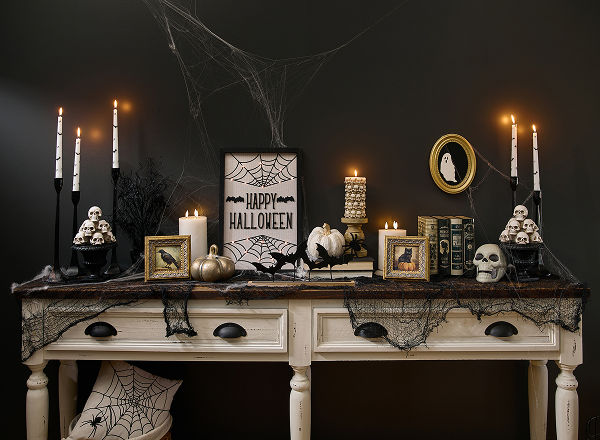 A spooky Halloween-themed foyer table is decorated with classic Halloween decorations from pOpshelf featuring skeletons, Halloween skulls, moody gothic-inspired framed art, bat candlesticks, a spider web tablecloth, brass pumpkins, and more affordable Halloween themed finds.