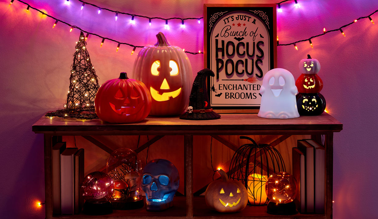 Halloewen string lights and light-up witch's hat, wicker pumpkin, jack-o-lanterns, ghosts & more on an entryway table.