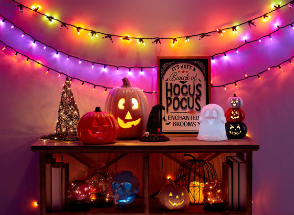 Halloewen string lights and light-up witch's hat, wicker pumpkin, jack-o-lanterns, ghosts & more on an entryway table.