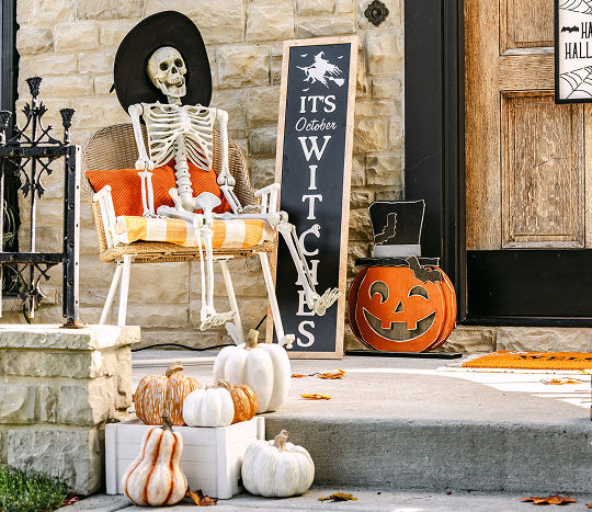 A stone home’s front porch is decorated for Halloween with affordable Halloween decor from pOpshelf, featuring a full-sized decorative skeleton wearing a witch hat sitting in an outdoor chair, various pumpkin accessories, and a front porch leaner that says “It’s October Witches.”
