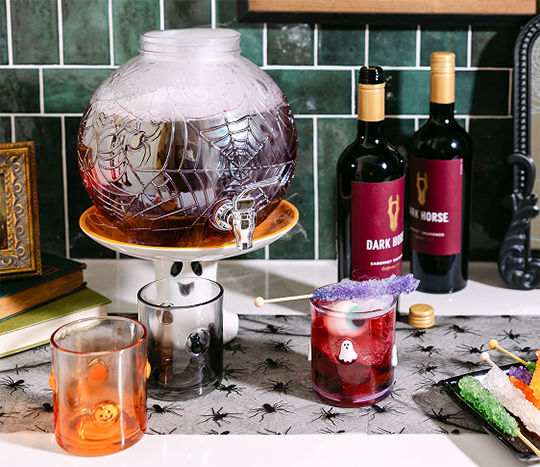 A kitchen counterspace is decorated with a Halloween spider-themed drink dispenser for parties and featured sitting on top of a ghost-themed riser with pumpkin and Halloween icon glasses from pOpshelf.