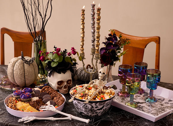 Spooky party table with black & white skeleton and bat serveware, skull goblets, candles, and decor, skeleton tongs & more.