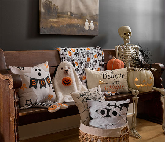 A wooden bench features Halloween pillows and a throw blanket from pOpshelf featuring ghosts, mummy, spider, and Halloween themed pillows with a lifesize skeleton sitting among them.