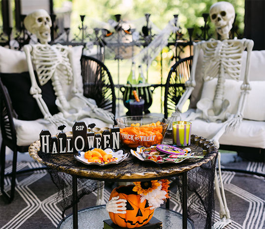 A porch is set with two faux skeletons sitting in lounge chairs with a table decorated with Halloween candy and snacks and decorated with fall florals and a classic Halloween Jack-O-Lantern themed pumpkin.
