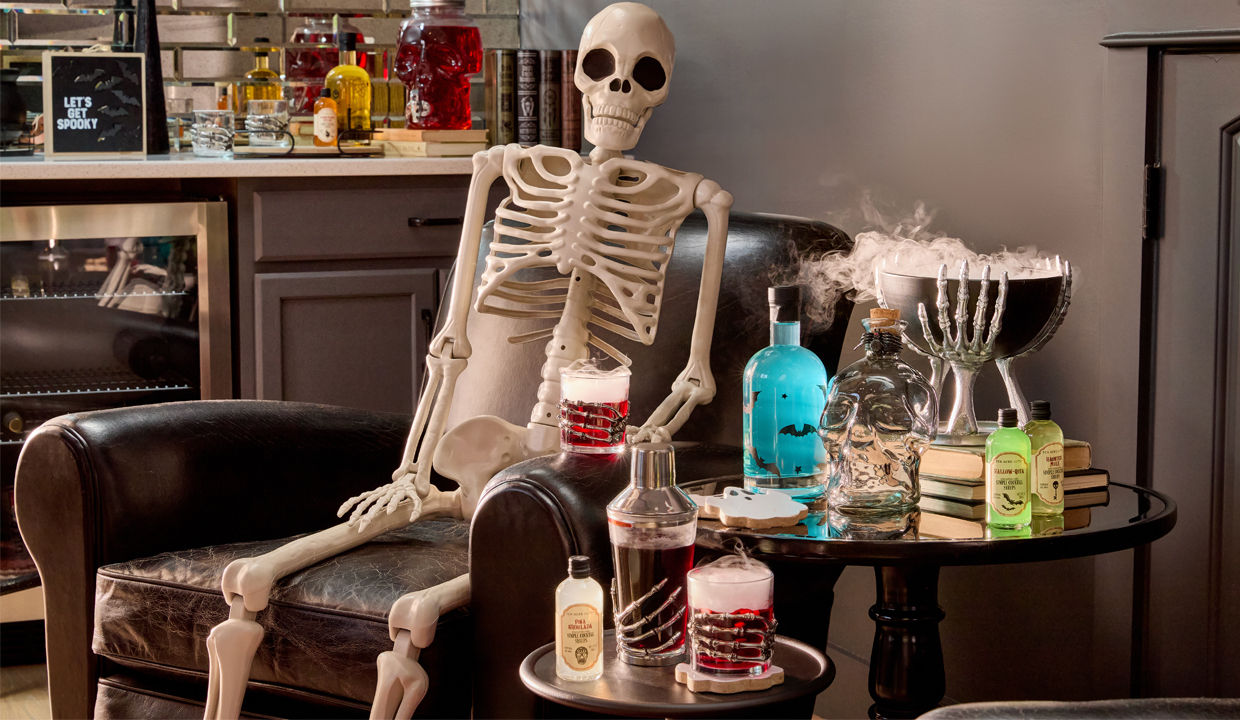 Life-size skeleton and skeleton hand cocktail glasses, shaker, and decorative bowl with cocktail mixers and Halloween decor.