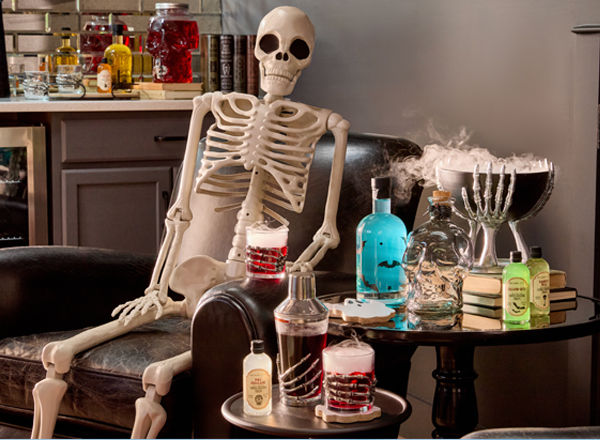 Life-size skeleton and skeleton hand cocktail glasses, shaker, and decorative bowl with cocktail mixers and Halloween decor.