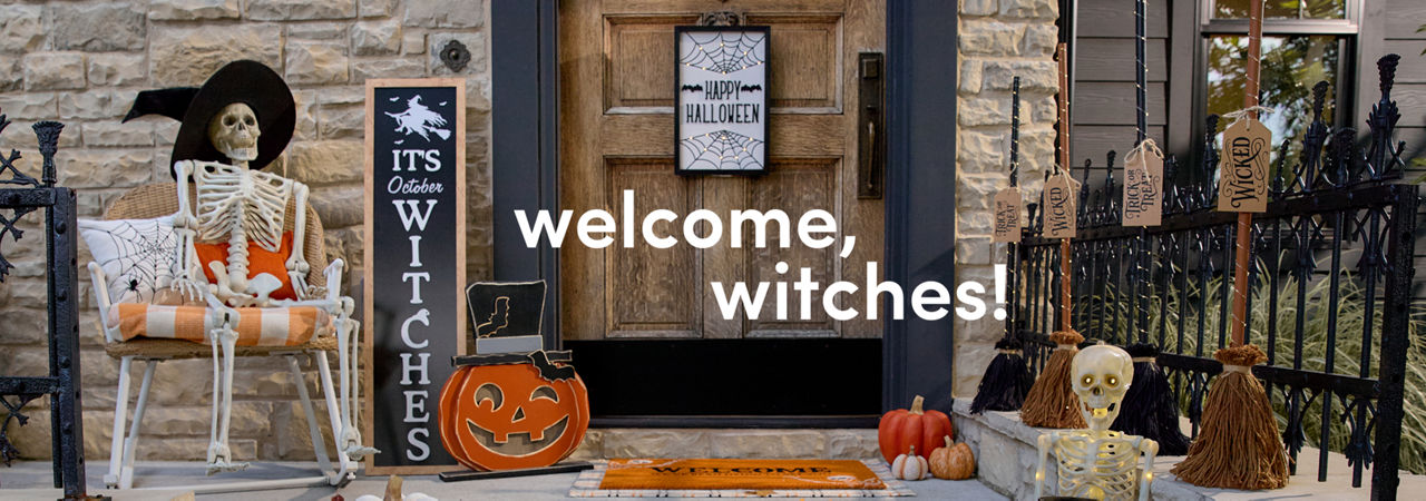 A stone home’s front porch is decorated for Halloween with affordable Halloween decor from pOpshelf, featuring full-sized decorative skeletons, a decorative candy cauldron, various pumpkin accessories, and a front porch leaner that says “It’s October Witches.”
