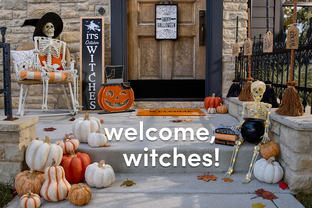 A stone home’s front porch is decorated for Halloween with affordable Halloween decor from pOpshelf, featuring full-sized decorative skeletons, a decorative candy cauldron, various pumpkin accessories, and a front porch leaner that says “It’s October Witches.”