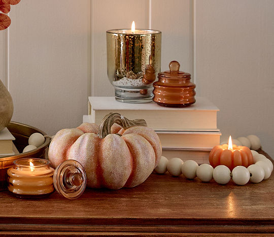 Candles are displayed with fall aesthetic decor including pumpkins and stacked books in an autumn color palette on a dark wooden table.
