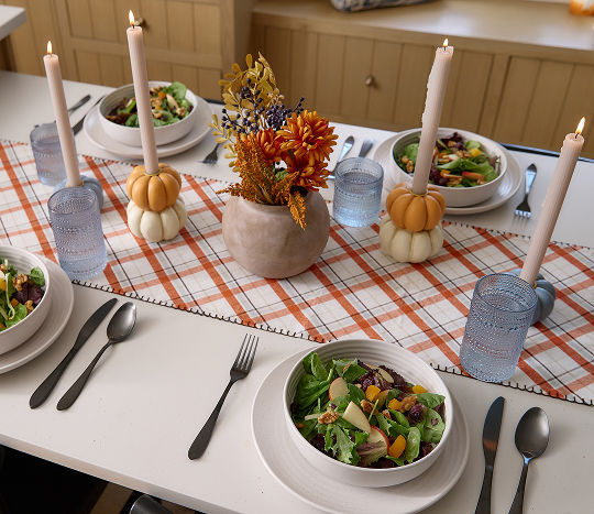 An autumn themed tablescape decorated with simple white serveware and paired with warm-toned fall decor including orange plaid print table runner, pumpkin candles, and a seasonal autumn floral arrangement.