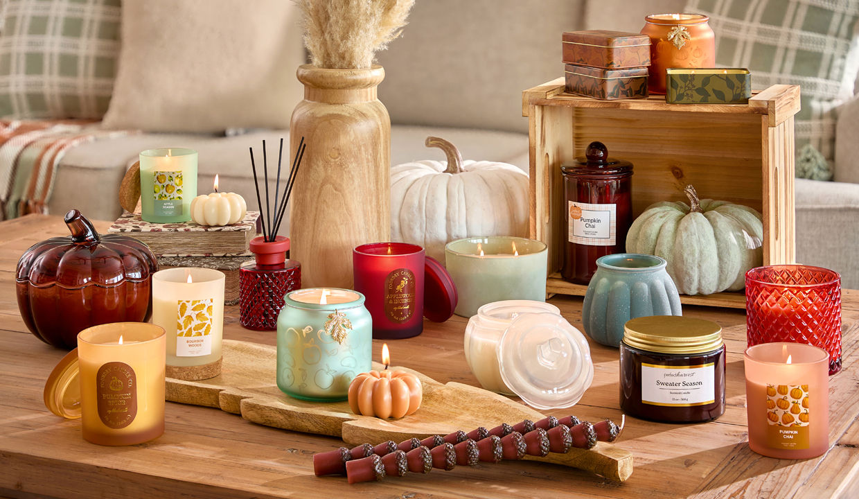 Collection of fall container and jar candles in various colors, fall scents, and shapes in front of a sofa with fall pillows.