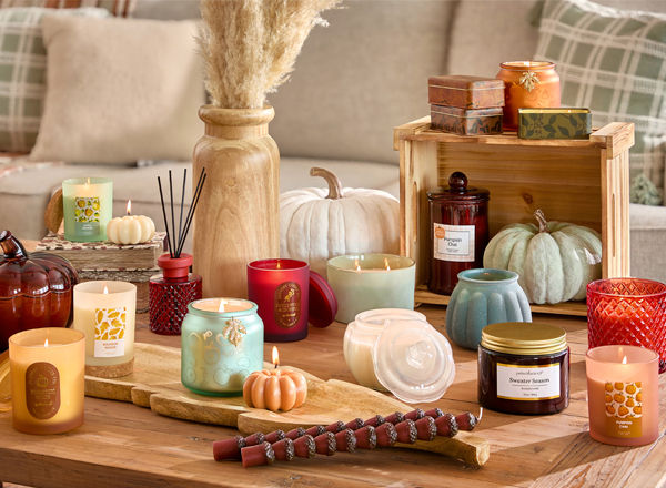 Collection of fall container and jar candles in various colors, fall scents, and shapes in front of a sofa with fall pillows.