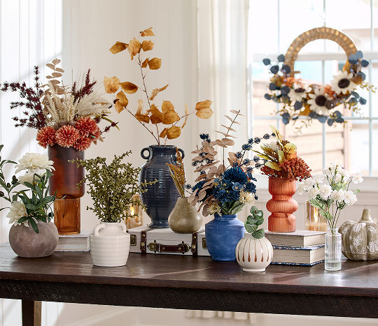 A dark wood table displays a warm-toned assortment of fall floral arrangements in a variety of vases consisting of various shapes and sizes.