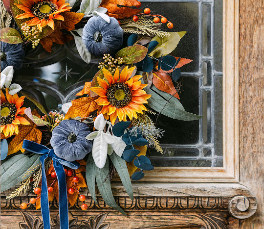 A rustic wooden front door displays a faux wreath made with aesthetic fall leaves, florals, and mini blue pumpkins.