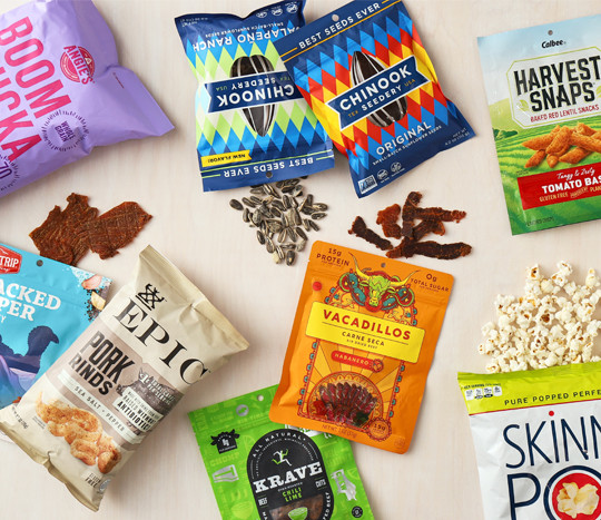 Healthier snack options on a table: Boom Chicka Pop, Epic Pork Rinds, Skinny Pop, Krave Jerky, Harvest Snaps, Chinook Seeds & more.