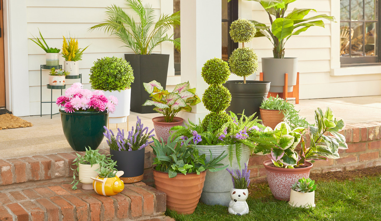 Various colors and designs of pOpshelf outdoor planters, flower pots, and plant stands filled with artificial greenery and flowers on a front porch.