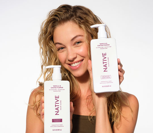 A woman with loose natural curls holding large bottles of Native haircare from pOpshelf.