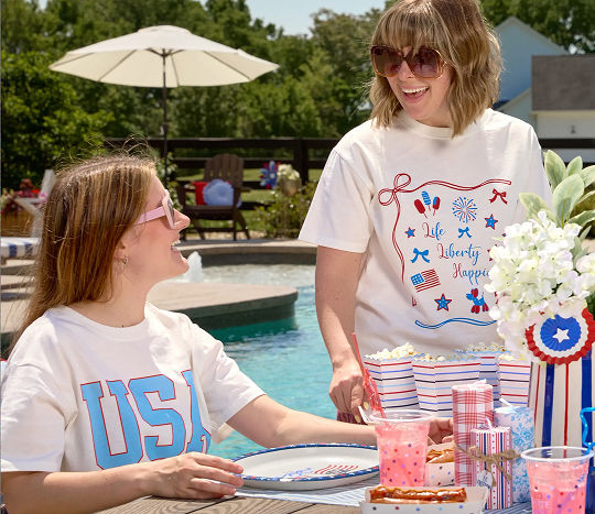 Two smiling women gather at a poolside dining table wearing patriotic apparel from pOpshelf.