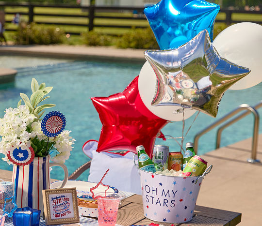 Star-shaped balloons in red, white & blue color options float poolside from a bucket of drinks displayed in an ice bucket.