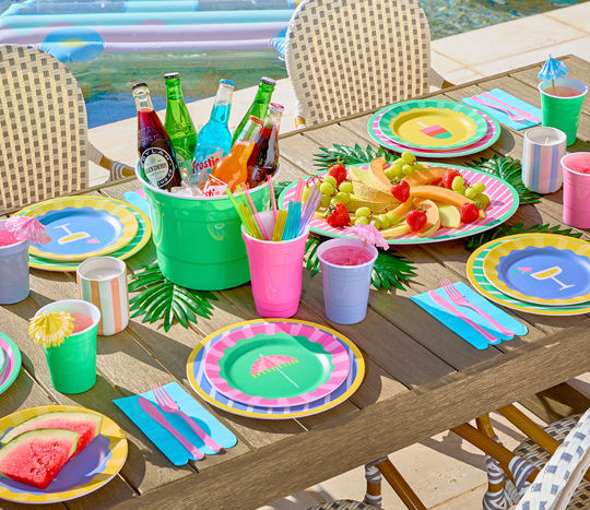 Poolside table set with colorful melamine dinnerware with stripes and summer icons, ice bucket, and matching drinkware.