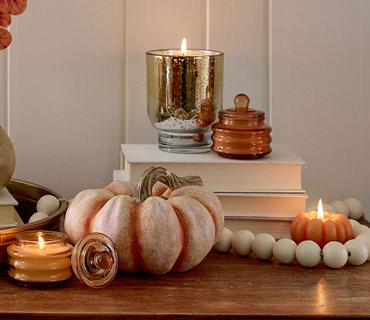 Candles are displayed with fall aesthetic decor including pumpkins and stacked books in an autumn color palette on a dark wooden table.