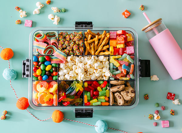 A snackle box from pOpshelf is filled with various snacks including gummy treats, popcorn, pretzels, and more with a water bottle featured lying beside the box.