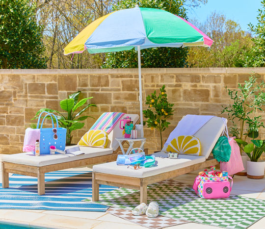 Lounge chairs with towels, pillows, white side table, rainbow umbrella, cooler, tote bags & accessories.