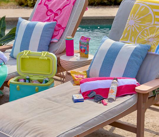 Poolside lounge chairs are decorated with cozy pillows, soft towels, and pool day essentials from pOpshelf including sunscreen, sunglasses, a cooler, and snacks.