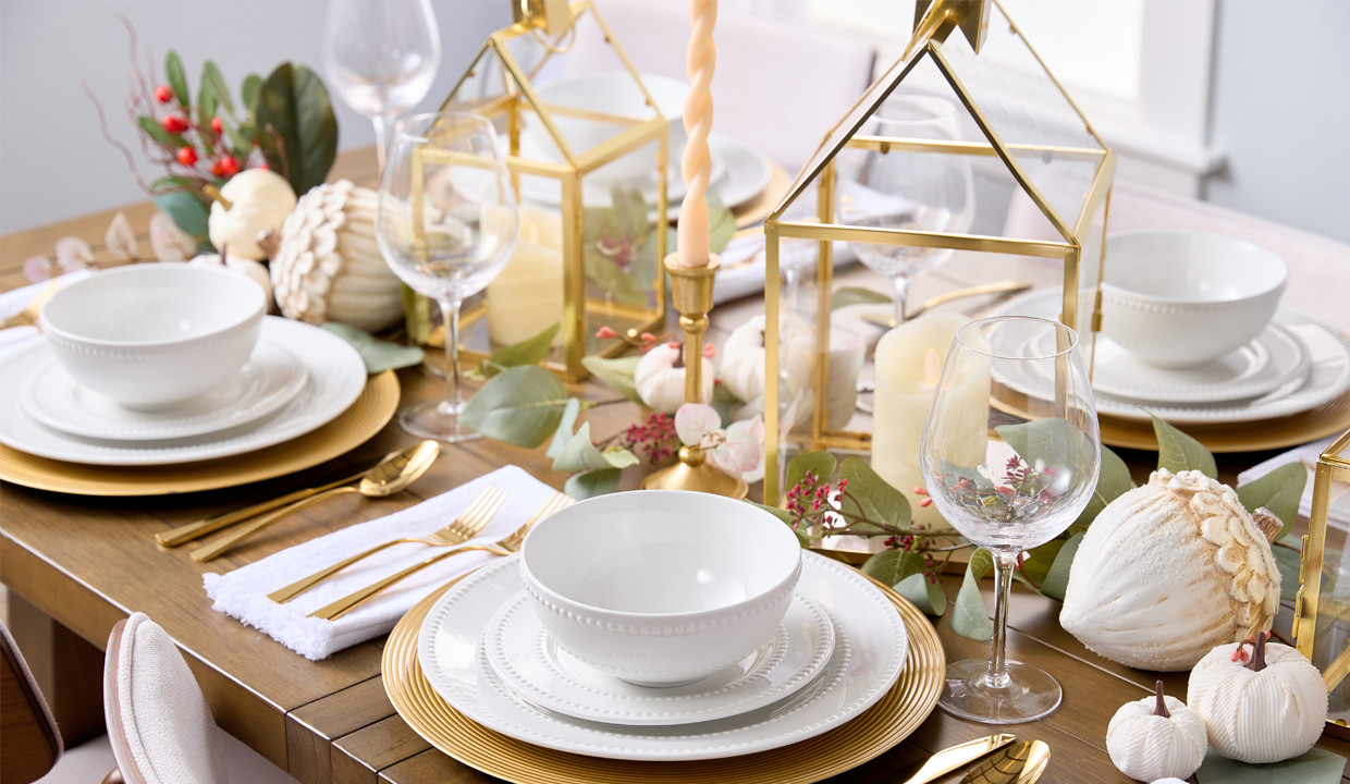 Elegant Thanksgiving tablescape: white dinnerware gold chargers & flatware, centrepiece with white pumpkins, large wooden acorns, gold-framed house lanterns, and faux greenery.