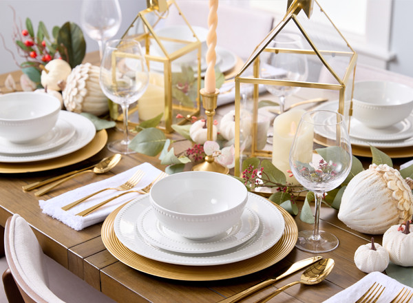 Elegant Thanksgiving tablescape: white dinnerware gold chargers & flatware, centrepiece with white pumpkins, large wooden acorns, gold-framed house lanterns, and faux greenery.