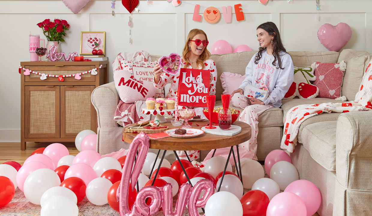 Galentine's Day living room setup, girls on couch toasting with heart-shaped plastic champagne glasses drinks, surrounded by Valentine's decor, balloons, pillows, throws, food, garland, banners, and more.