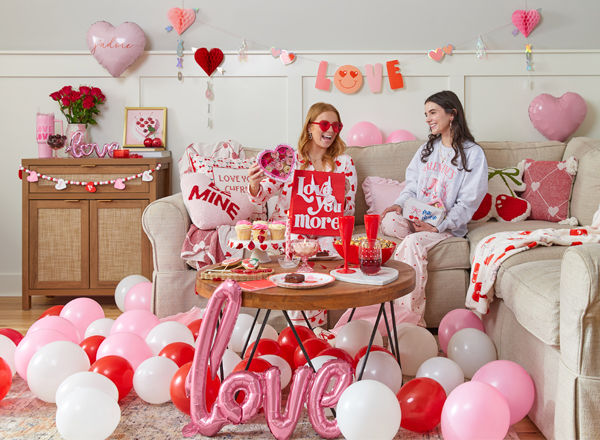 Galentine's Day living room setup, girls on couch toasting with heart-shaped plastic champagne glasses drinks, surrounded by Valentine's decor, balloons, pillows, throws, food, garland, banners, and more.