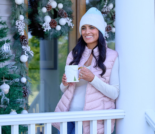 Woman in pink puffer vest and cream and blue hat on the front porch drinking coffee.