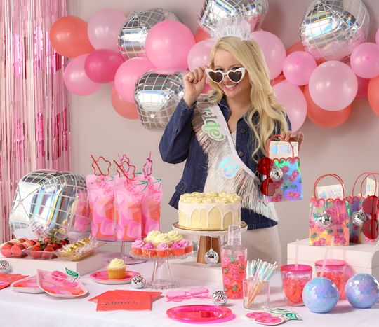 Bachelorette party table with pink and silver disco party supplies: bride sash, sunglasses, and tiara; party plates, napkins, utensils, party balloon and fringe decorations, favor bags and sunglasses, and more.