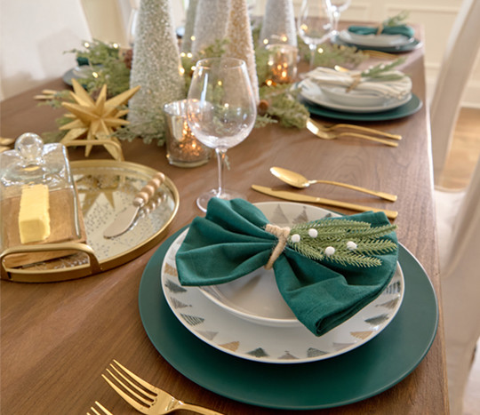 Dining table set for Christmas with ceramic Christmas tree designed dinner plates and bowls, wine glasses, solid and green-striped napkins with pine napkin holders, cone tree centerpiece with faux greenery, lights, and large sparkly tabletop decor.