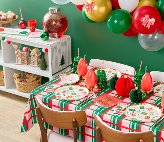 Christmas party table with festive gift and Santa themed party plates and napkins, ornament and glass tree cups with straws, honeycomb ornament centerpiece, and plastic ornament drink dispenser.