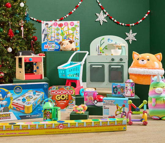 Toys in front of Christmas tree: Sonic air hockey table, wooden kitchen & tool bench sets, play shopping cart, play-doh, plush, games & more.