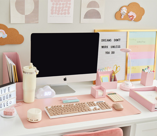 Work or study desk outfitted with neutral-toned desk accessories such as a white keyboard with pink keys, a pink mouse, calculator & desk organizers, markers, pens, gold scissors & more.