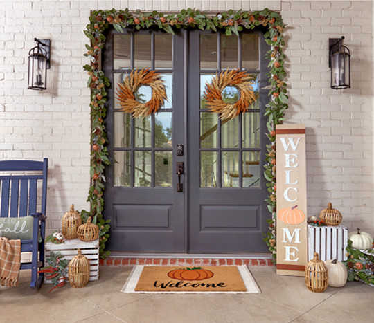 Front porch decorated with fall garland, fall wreaths, "welcome" porch leaner, wicker lantern pumpkins, fall pillow & throw, fall coir mat, and fall flowers.