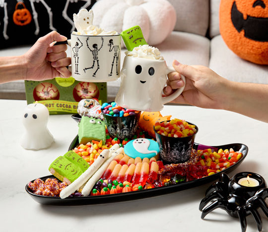 Halloween candy charcuterie on a black witch hat platter with Halloween mugs, spider and ghost candles, and cocoa bombs.