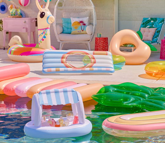 Colorful inflatable pool floats, swim rings, and rafts from pOpshelf in various designs: llama, peach, camera, palm leaf, floating buffet, and more.