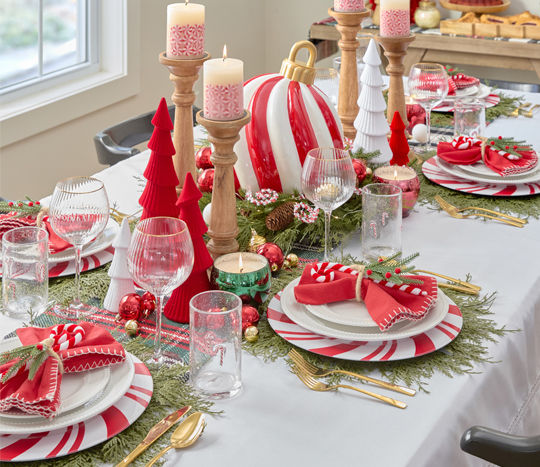 Dining table set for Christmas with peppermint candy charger plates and white beaded dinnerware, faux pine placemats, candycane water goblets, and a giant peppermint swirl ornament centerpiece with red and green trees, candles & more.
