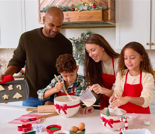 Family baking cookies in Christmas mixing bowls and more in a kitchen decorated for Christmas.