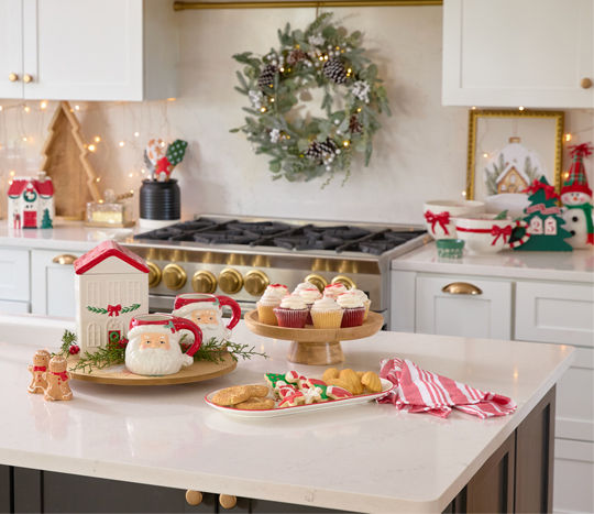 Christmas mixing bowl, wooden cake stand and lazy susan, tree-shaped charcuterie tray, village house cookie jar, Santa mugs, gingerbread man salt & pepper shakers, and more in a kitchen decorated for Christmas.